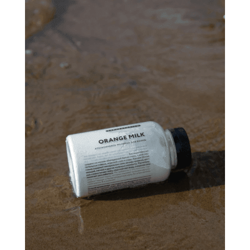 Plastic bottle labeled 'Orange Milk' resting on sandy beach with water.